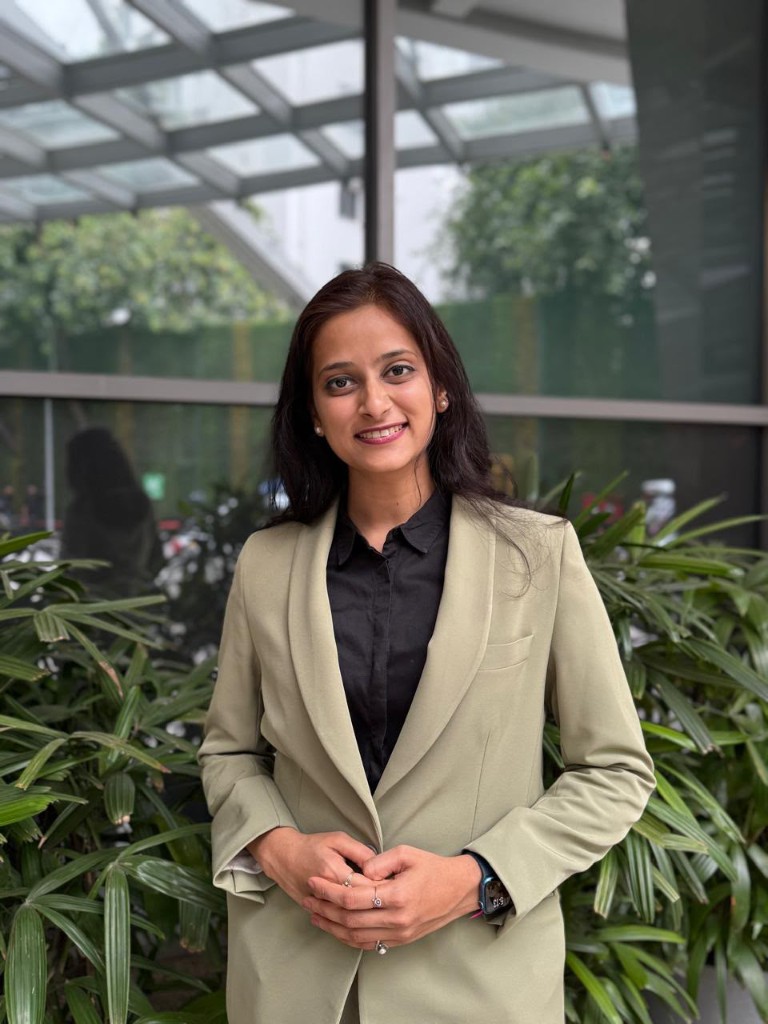 Portrait of the founder, a woman in a sage-green blazer standing among green plants.
